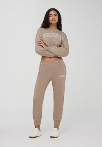 Felpa beige con la scritta "PHOENIX" stampata sulla parte anteriore, abbinata a pantaloni della tuta a gamba affusolata, entrambi in morbido tessuto e design casual.