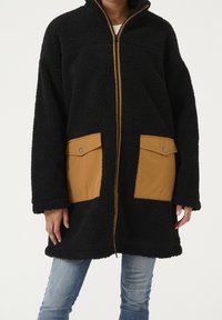 Svart fleecejacka med bruna framlappsfickor och dragkedja, bärs över ljusblå jeans av en stående person.