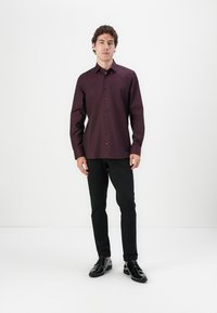 Burgunderfarbendes Langarmhemd mit Knopfleiste, dunklen Knöpfen und einer glatten Textur, kombiniert mit schwarzen Hosen und glänzenden schwarzen Schuhen.