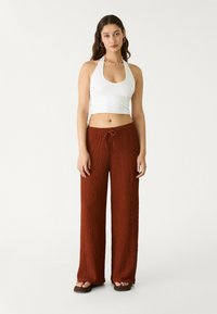Bílý zkrácený halter top a široké kalhoty v barvě rezavého textilu s šňůrkou. Ploché hnědé sandály. Jednoduchý náhrdelník s korálky.