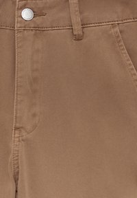 Pantalon en denim marron en gros plan montrant le bouton, la boucle de ceinture, la poche avant et la texture du tissu.