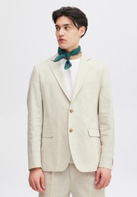 Ljusbeige linneblazer med kavajklaffar, två framfickor och ljusa knappar, parat med en vit t-shirt och en grönmönstrad scarf.