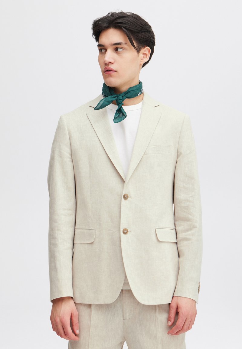 Ljusbeige linneblazer med kavajklaffar, två framfickor och ljusa knappar, parat med en vit t-shirt och en grönmönstrad scarf.