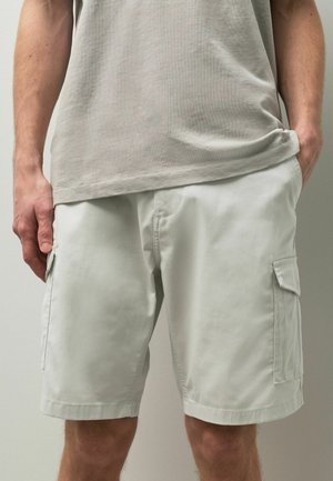 Mann trägt hellbeige Cargo-Shorts mit Seitentaschen, eine Hand in der Tasche, und ein beigefarbenes T-Shirt, das teilweise in die Hose gesteckt ist.