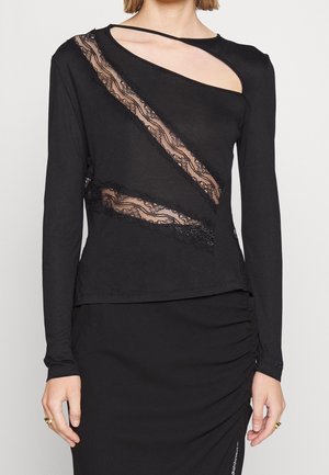 Femme portant un haut noir à manches longues avec des découpes en dentelle diagonales et un col asymétrique, associé à une jupe noire froncée.