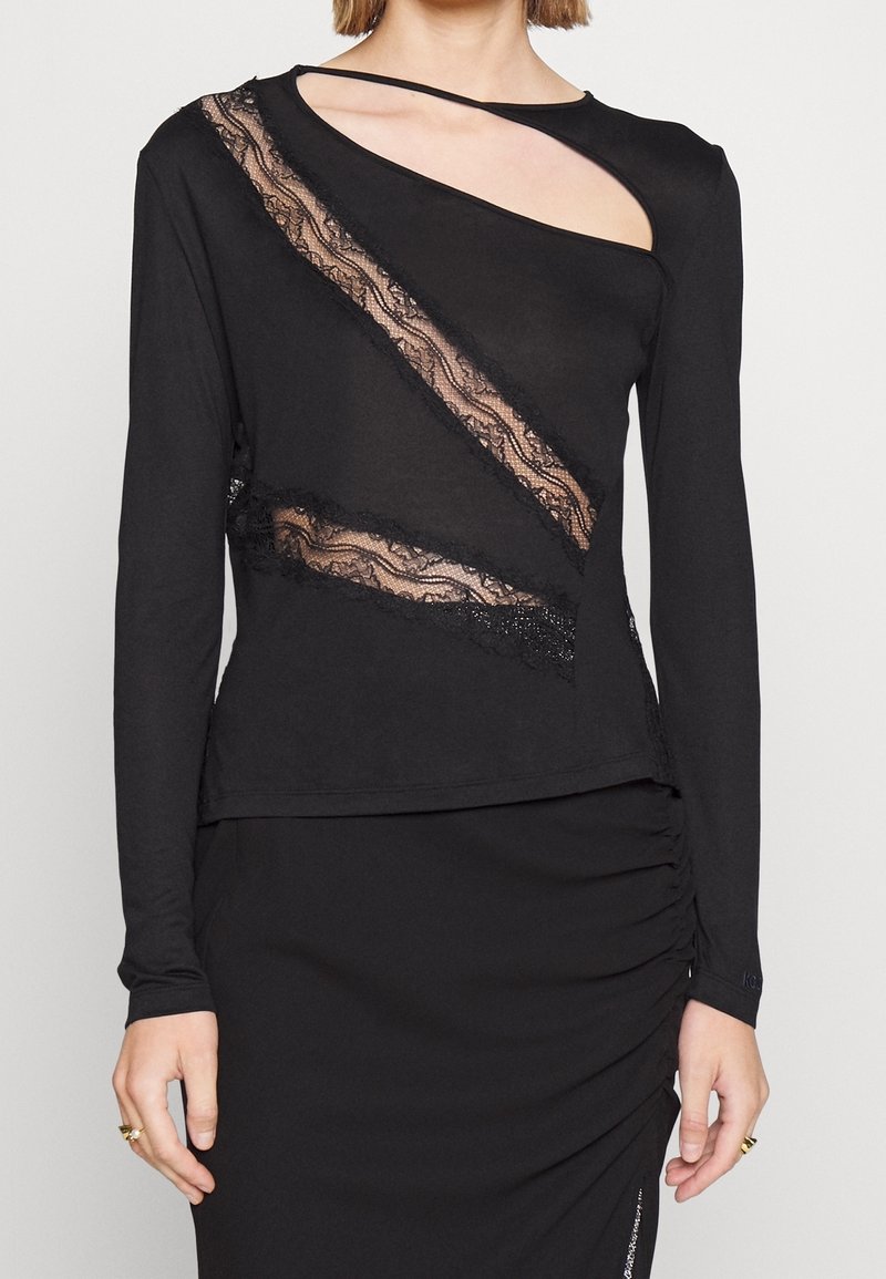 Femme portant un haut noir à manches longues avec des découpes en dentelle diagonales et un col asymétrique, associé à une jupe noire froncée.