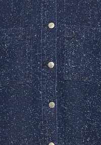 Tissu en denim bleu foncé tacheté avec fermeture verticale à boutons et deux poches poitrine sécurisées par des boutons-pression en métal argenté.