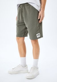 Mand med olivengrønne trækbåndshorts med "Premium Quality" mærke, hvide ankelstrømper og hvide sneakers foran en ensfarvet baggrund.