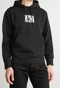 Persona vistiendo una sudadera negra de Calvin Klein con bolsillo frontal y cordones, combinada con pantalones negros, frente a un fondo claro y liso.