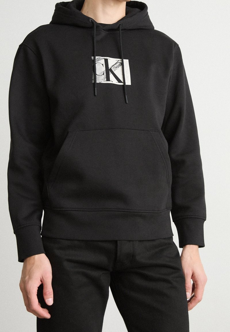 Persona vistiendo una sudadera negra de Calvin Klein con bolsillo frontal y cordones, combinada con pantalones negros, frente a un fondo claro y liso.