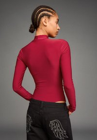 Crvena bodysuit s dugim rukavima u kombinaciji s crnim trapericama koje imaju dizajn krila obrubljen rhinestones-ima. Glatka tekstura, visoki ovratnik.
