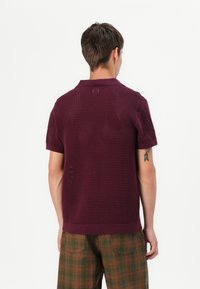Polo en maille bordeaux à manches courtes et motif texturé. Associé à un pantalon à carreaux marron avec des accents verts.