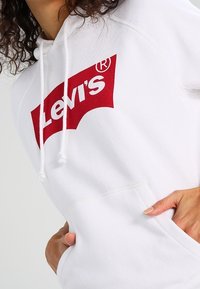 Sudadera blanca con el logo rojo de "Levi's"; cuenta con cordones, un bolsillo canguro y puños acanalados para un ajuste cómodo.