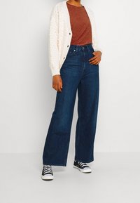 Jeans en denim bleu foncé à jambes larges, associés à un haut texturé couleur rouille et un cardigan en tricot crème avec des détails de boutons. Des baskets noires complètent la tenue.