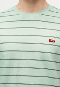 Ανοιχτό πράσινο μπλουζάκι με στρογγυλή λαιμόκοψη, λεπτές οριζόντιες μαύρες ρίγες και ένα μικρό κόκκινο έμβλημα με το λογότυπο της Levi’s στο αριστερό στήθος.