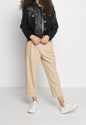 Personne portant une veste en cuir noire, un pantalon beige raccourci et des baskets blanches, debout les mains jointes devant un fond uni.