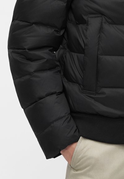 Gros plan d'une personne portant une doudoune noire avec une main dans la poche d'un pantalon beige clair.