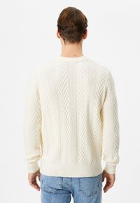 Suéter de crema grueso con patrón de trenzado texturizado, puños y dobladillo acanalados, cuello redondo y ajuste relajado, combinado con jeans de color azul claro.