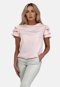 Jasnoróżowa bluzka z krótkim rękawem, z falbanami na rękawach, z miękkiego materiału, okrągły dekolt, luźny krój, zestawiona z jasnymi spodniami.