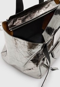 Metallisk sølv tote bag med en tekstureret overflade, sort inderside, lynlåslukning og kontrasterende sorte stropper. Har en sidelomme.