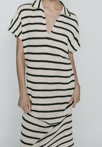 Una donna che indossa un vestito corto a maniche corte e scollatura a V, con righe orizzontali bianche e nere, in piedi con le mani rilassate ai lati.