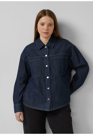 Młoda kobieta o prostych, jasnobrązowych włosach, ubrana w ciemnoniebieską dżinsową koszulę na guziki i czarne spodnie, stojąca na tle jednolitego jasnoszarego tła.