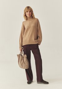 Maglione beige a maglia con colletto alto, maniche ampie e orlo a coste, abbinato a pantaloni viola scuro e a una morbida borsa a tracolla beige.