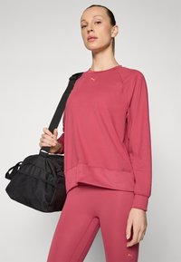 Sweatshirt rosa con orlo a costine e scollatura a girocollo, abbinato a leggins coordinati; tiene in mano una borsa a tracolla nera con dettagli in rete.