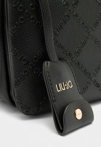 Schwarze Ledertasche mit geprägtem Logo-Design, strukturiertem Finish und goldfarbener Hardware-Akzent am Tragegurt.
