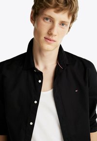 Chemise noire à boutons avec un col pointu, ornée de boutons blancs et d'un petit logo rouge, blanc et bleu sur la poitrine. T-shirt blanc visible en dessous.