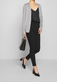 Cardigan gris sur un haut noir à col en V, associé à un jean skinny noir et des chaussures à talons carrés en cuir verni noir. Tient un petit sac à main noir.