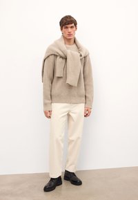 Beež ribatud sviiter, millele on kantud sobiv sall õlgadel, koos kreemivärvi pükste ja mustade kingadega, neutraalse tausta taustal.