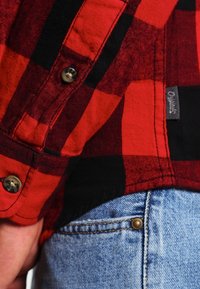 Camisa de franela a cuadros rojos y negros con botones de metal y una etiqueta de logo, combinada con unos jeans de mezclilla azul claro que tienen un detalle de remache de latón.