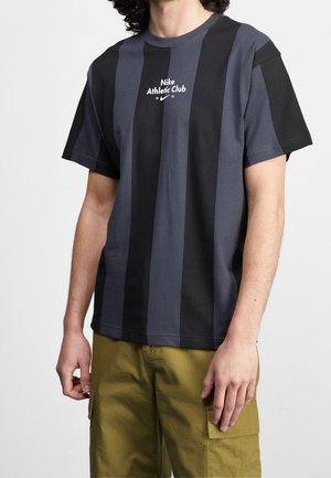 Man draagt een zwart en donkergrijs verticale gestreepte Nike Athletic Club T-shirt en olijfgroene cargoshorts, staand tegen een effen achtergrond.