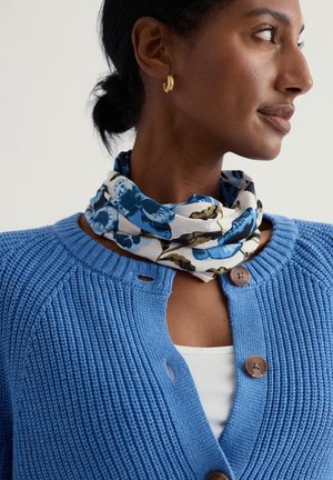 Blauwe gebreide cardigan met een ronde halslijn en bruine knopen, gecombineerd met een wit katoenen topje. De sjaal is bedrukt met een bloemmotief in blauw en groen.