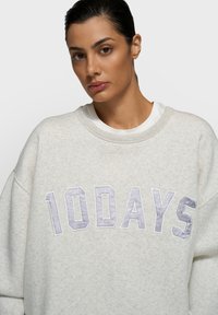 Sweat-shirt oversize gris clair avec "10DAYS" brodé en lettres capitales violet pâle sur le devant. Tissu doux et texturé avec un col côtelé.