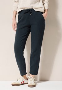 Zwarte, textuurachtige broeken met een relaxte pasvorm, voorzien van een elastische tailleband en trekkoord. Gecombineerd met witte en beige sneakers.