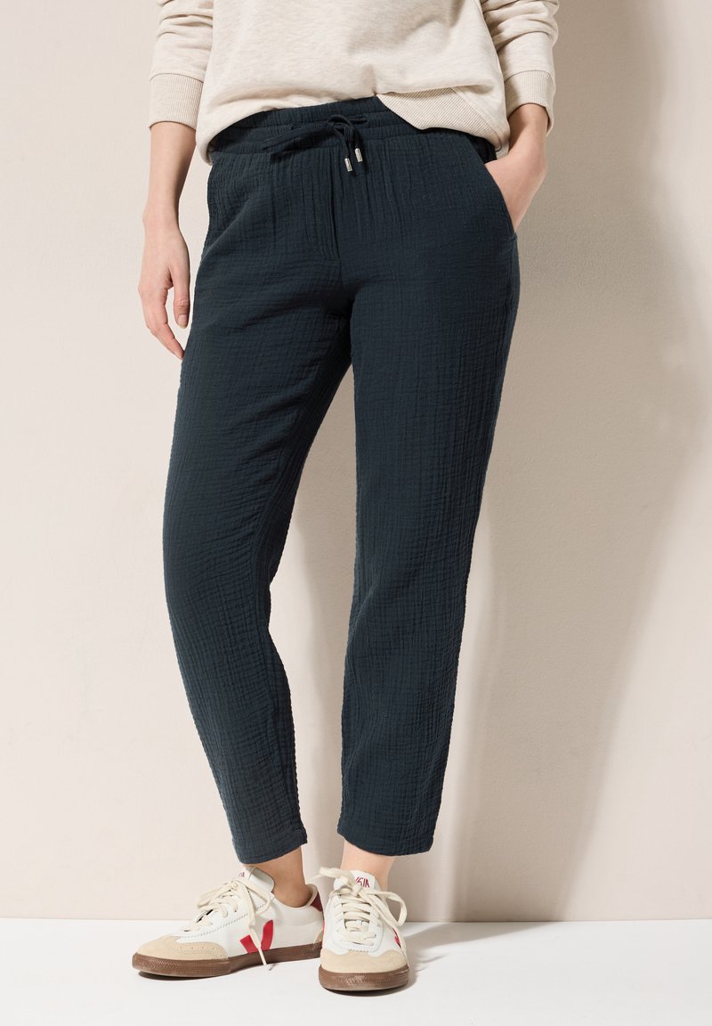 Zwarte, textuurachtige broeken met een relaxte pasvorm, voorzien van een elastische tailleband en trekkoord. Gecombineerd met witte en beige sneakers.
