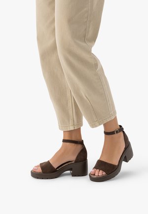 Stopy w brązowych zamszowych sandałach na słupku z odkrytymi palcami i paskami wokół kostek, w połączeniu z beżowymi, skróconymi spodniami na białym tle.