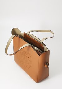 Sac à main en cuir beige avec poignées beige, fermeture éclair ouverte en haut et logo circulaire perforé à l’avant sur fond blanc.
