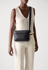 Schwarze Leder-Crossbody-Tasche mit strukturierter Oberfläche, ausgestattet mit einer vorderen Reißverschlusstasche und einem verstellbaren Tragegurt. Getragen über einem cremefarbenen Strickoberteil und hellen Hosen.