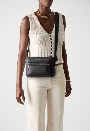 Borsa a tracolla nera in pelle con finitura testurizzata, dotata di una tasca anteriore con zip e una tracolla regolabile. Indossata sopra un top in maglia crema e pantaloni chiari.