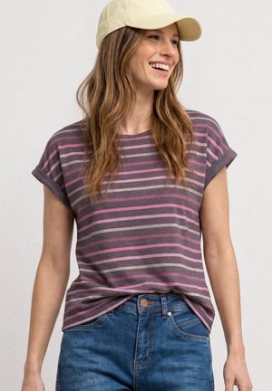 Femme souriante regardant vers la droite, portant une casquette beige, une chemise rayée mauve et rose à manches courtes, et un jean bleu avec le devant rentré.