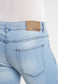 Vaqueros de denim azul claro con un diseño ajustado, que cuentan con un parche de cuero etiquetado con 'YOURTURN' en la cinturilla y un bolsillo trasero visible.