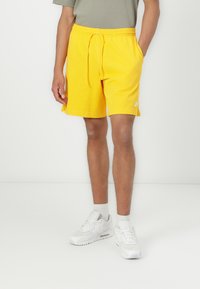 Shorts de sport jaunes en tissu doux, avec une taille élastique et un cordon de serrage, des fentes sur les côtés et un petit logo blanc.