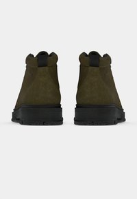 Pár olivovo zelených semišových kotníkových čižiem s čiernymi sťahovacími pútkami a hrubými čiernymi gumovými podrážkami, zobrazených zozadu na bielom pozadí.