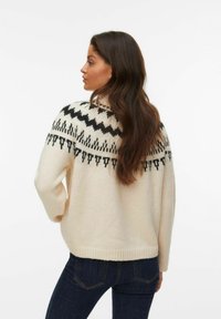 Cremfarbener Strickpullover mit einem schwarzen geometrischen Muster an der Schulterpartie. Das Design umfasst lange Ärmel und einen lockeren Schnitt, dargestellt von hinten.