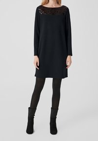 Nainen, jolla on yllään musta pitkähihaiset mekko, jossa on paljettiyksityiskohta, mustat sukkahousut ja mustat nilkkurit, seisoo yksinkertaista taustaa vasten.