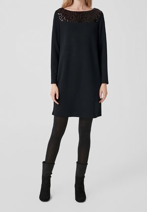 Donna che indossa un vestito lungo nero con yoke di paillettes, collant neri e stivaletti neri, in piedi davanti a uno sfondo neutro.