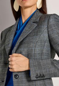 Grijze geruite blazer met blauwe lijnen, gestructureerde schouders en knoopmanchetten. Gelaagd bovenop een blauwe top, met een close-up van de hand op de revers.